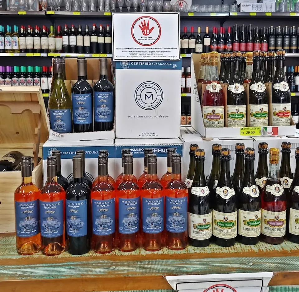 Mesa rústica com garrafas de vinho rosé, tinto e lambrusco, diante de prateleiras de vinhos e taças